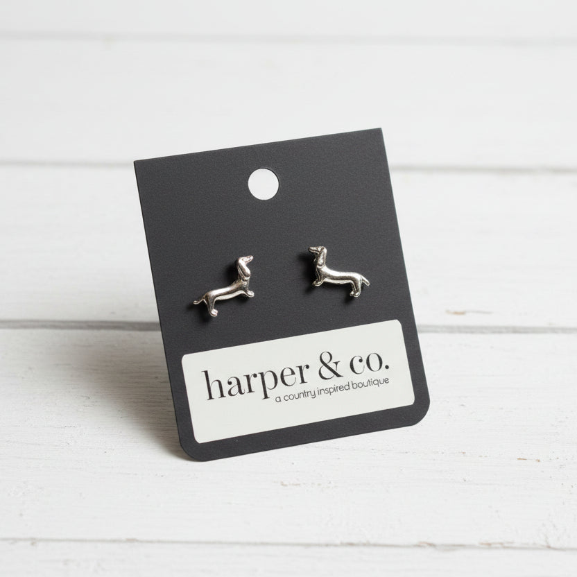 A pair of small silver stud earrings shaped like dachshunds, displayed on a black backing card labeled 'harper & co.' placed on a teal textured surface.