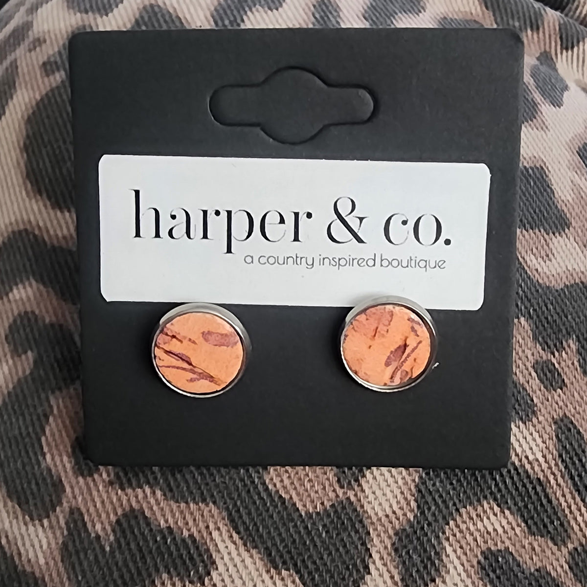 A pair of round copper-toned earrings displayed on a black card backing with the brand name 'harper & co.' printed on a leopard-print fabric background.