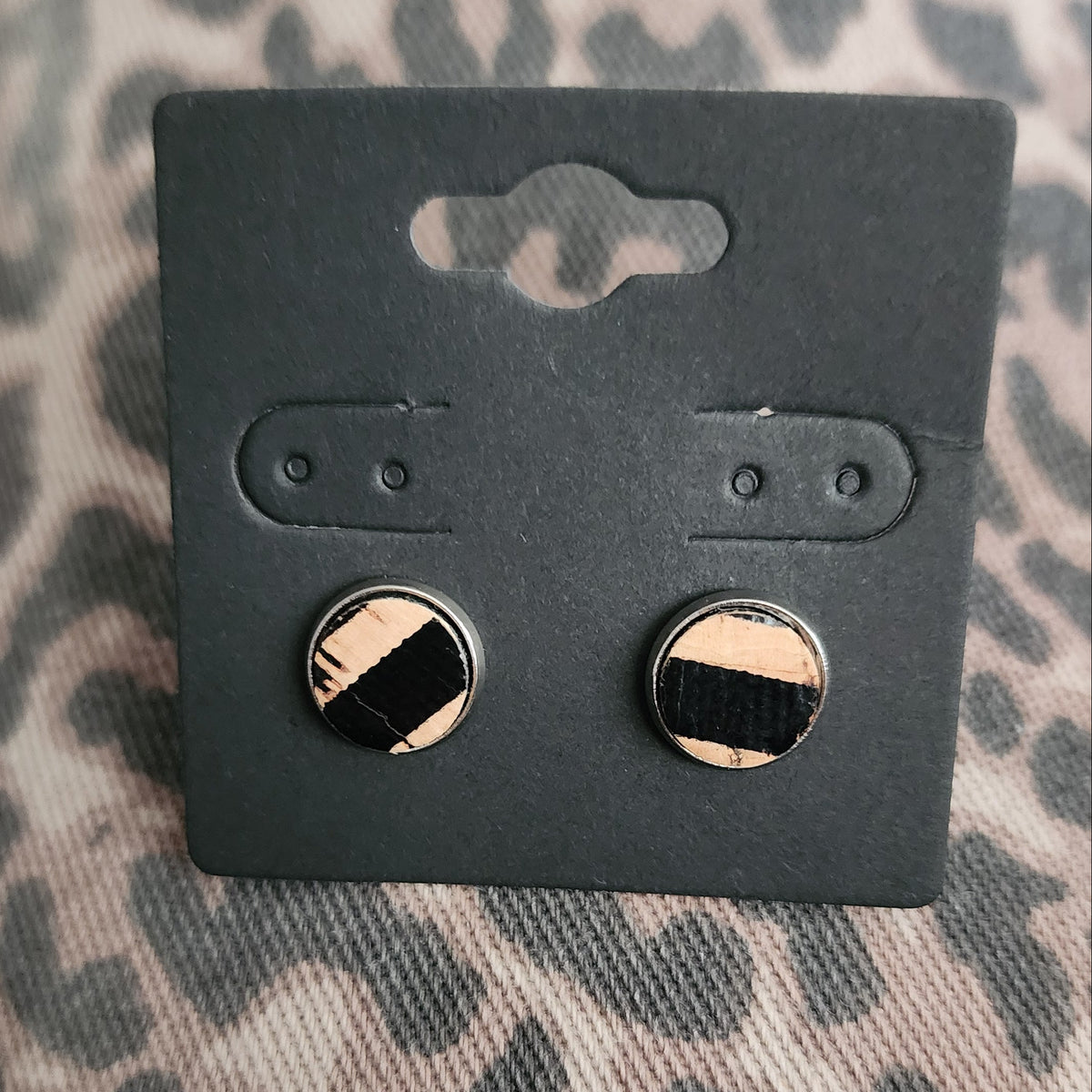A pair of round stud earrings with a black and khaki gold diagonal striped design, displayed on a black cardboard hanger against a leopard print fabric background.