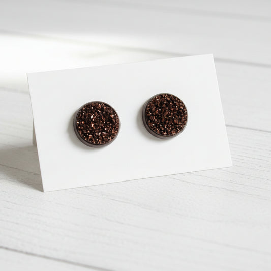 A pair of round brown stud earrings with a glittery silver and white speckled druzy finish, mounted on a white display card against a white background.