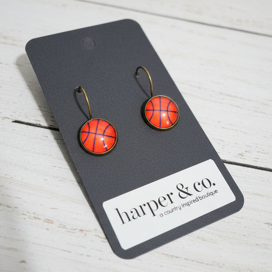 Pair of basketball earrings on a black card with 'harper & co.' branding against a white wooden background