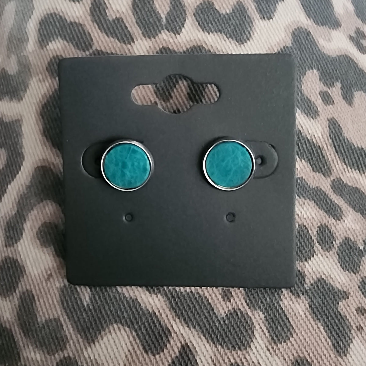 A pair of round stud earrings with turquoise-blue centers and silver-toned edges, displayed on a black backing card over a leopard-print fabric background.