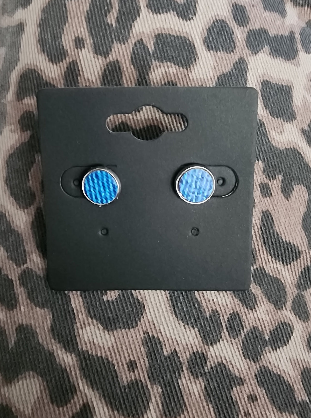 A pair of round stud earrings with a textured denim, mounted on a black display card, placed on a leopard print fabric background.