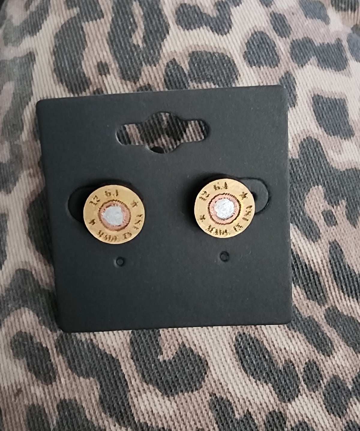 A pair of bullet-shaped stud earrings displayed on a black backing card, placed on a leopard-print fabric surface. The earrings have a gold-toned outer ring with a white center.