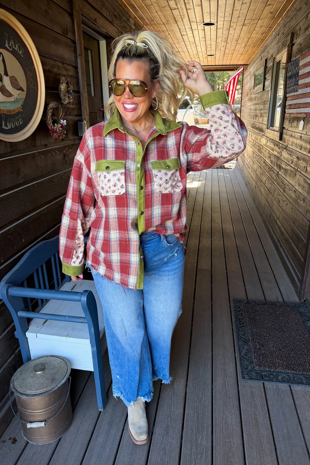 A woman stands on a rustic wooden porch wearing a red and white plaid button-up shirt with olive green and patterned accents, blue jeans, and tan boots.