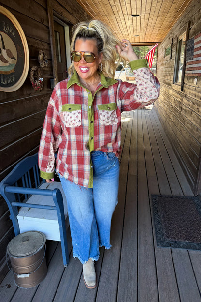 A woman stands on a rustic wooden porch wearing a red and white plaid button-up shirt with olive green and patterned accents, blue jeans, and tan boots.