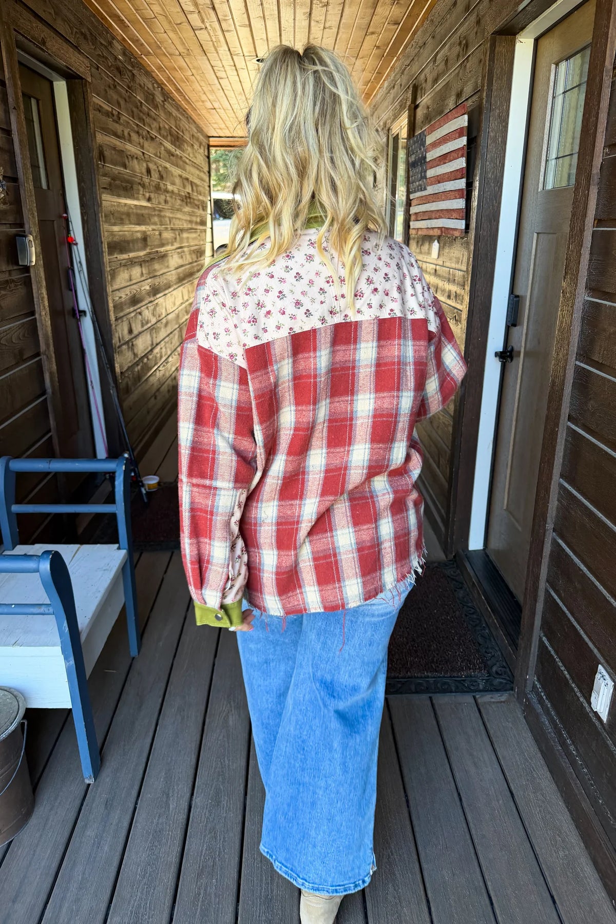 A woman viewed from behind wearing a red and white plaid patchwork shirt with a floral yoke, blue jeans, and beige boots, standing on a rustic wooden porch.