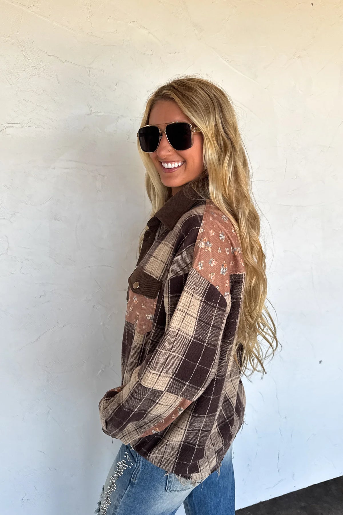 A woman with long blonde hair wearing a brown and beige plaid patchwork flannel shirt, blue jeans, and dark sunglasses, posing against a white stucco wall.