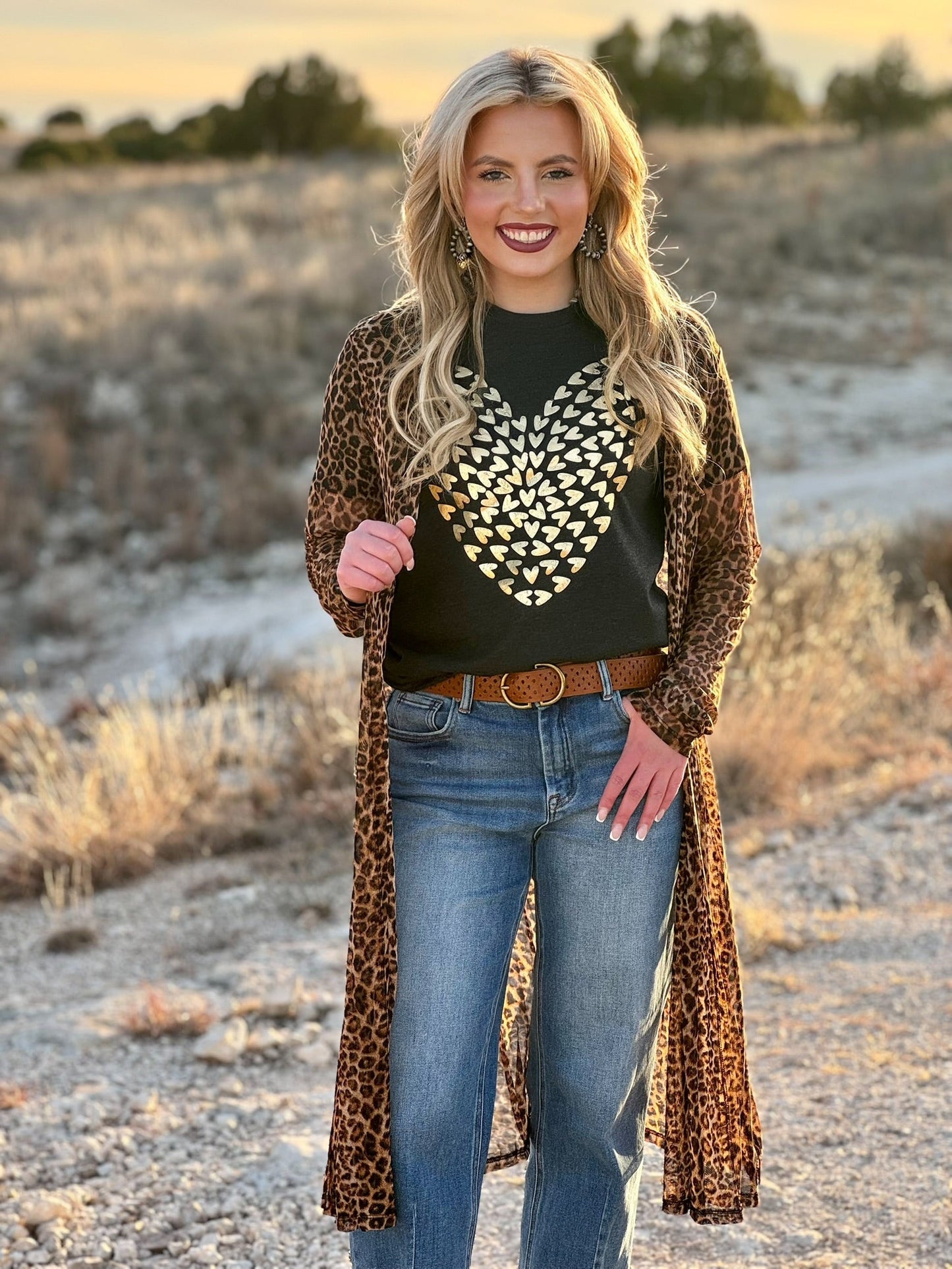 Woman standing in a desert landscape wearing a leopard print cardigan, black patterned shirt, and blue jeans.