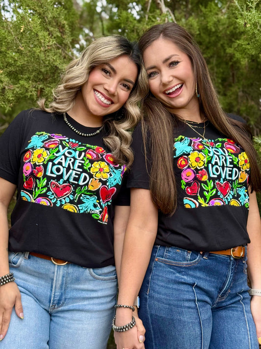 Two women wearing black t-shirts with colorful designs and text, standing outdoors.
