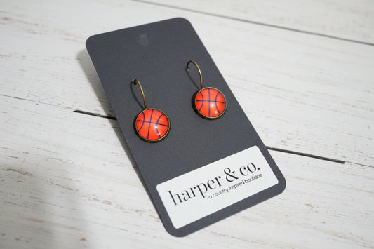 Pair of basketball earrings on a black card with 'harper & co.' branding against a white wooden background