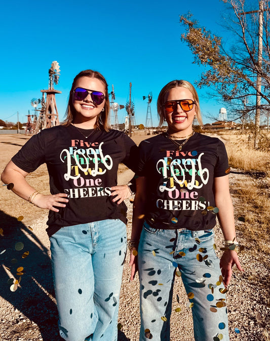 Two women wearing matching black t-shirts with colorful text in an outdoor setting.