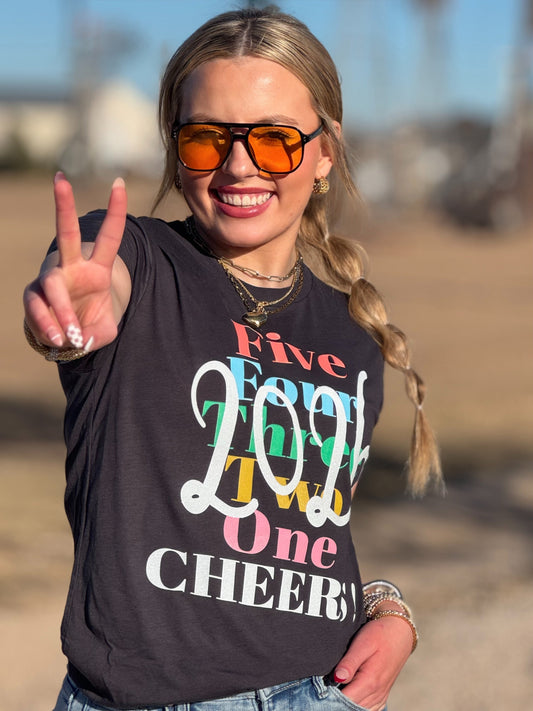 Person wearing a gray New Years Eve t-shirt with colorful text, making a peace sign outdoors.