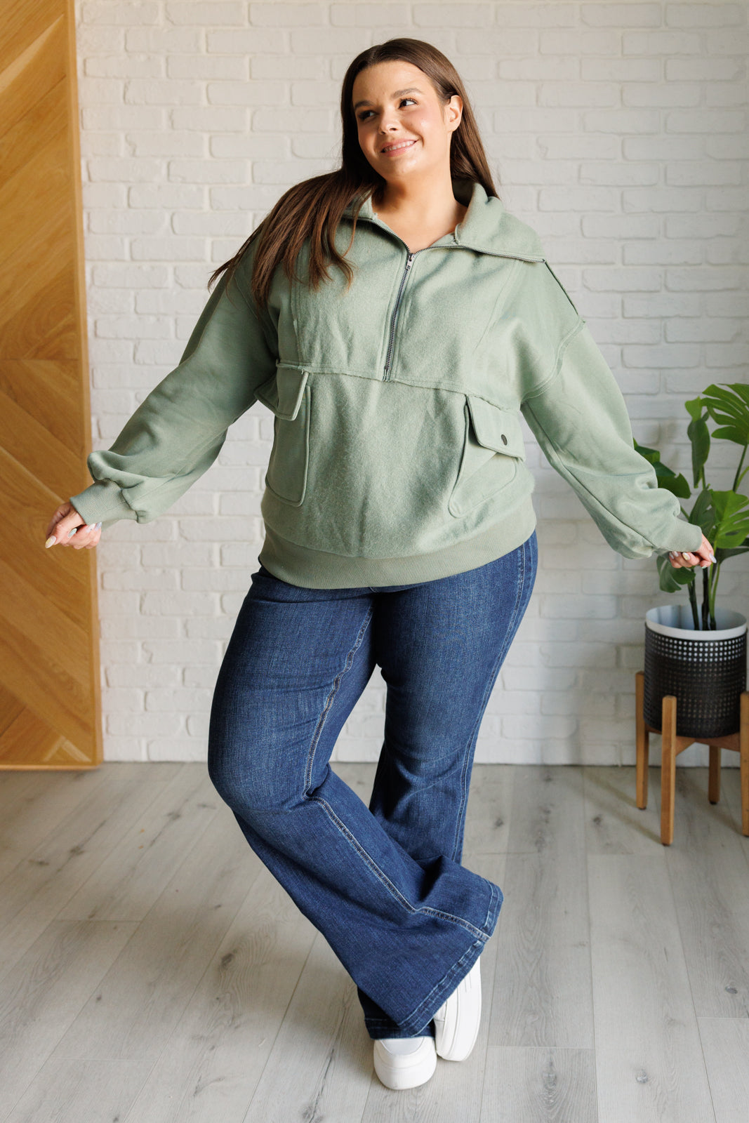 Woman wearing a green pullover and blue jeans standing indoors with a white brick wall background.